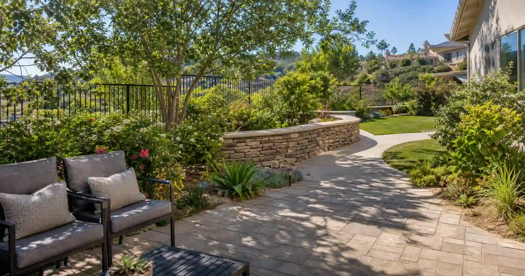 What is hardscaping? A backyard with a paver patio, curved walkway, and stacked stone retaining wall beside shrubs, flowers, and a tree